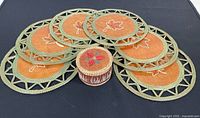 Six round trivets with maple leaf designs in a sweet grass woven frame arranged on black fabric.