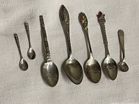 Photo showing all seven vintage sterling silver collectible spoons laid out on white fabric. Various designs visible including New York City, Empire State Building, and colored maple leaf motifs.