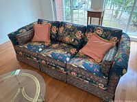 Front view of floral fabric sofa with peach gingham accent pillows on hardwood floor near window.