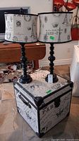 Two Paris-themed table lamps with black turned bases and fabric lampshades sitting on a matching small storage trunk.