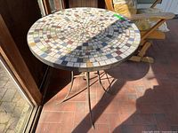 Image showing the round patio table with mosaic tile top and metal legs, with visible wear on the tile surface.
