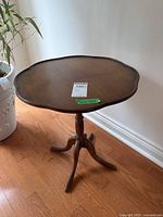 Full view of the vintage pie crust wooden table, showing top shape and pedestal base on hardwood floor next to plant.