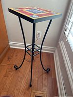 Tile top plant stand with colorful tiles and black metal legs standing on hardwood floor near an electrical outlet and window with white shutters.