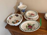 Photo showing the full set arranged on wooden surface: two serving bowls with fruit pattern, four dessert plates stacked, oval dish, and vase with plum design.