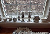 Photo showing the full set of Portmeirion Botanic Garden items including serving bowls, cream and sugar set, two candle holders, and candle warmer bowl lined up on a windowsill.