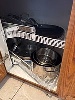 Two kitchen cabinet pullout shelves fully extended showing multiple stainless steel pots and pans, some with glass lids inside the cabinet.