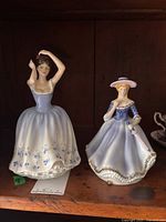 Two porcelain figurines on a wooden shelf: Royal Doulton Sheila HN2742 in white and lavender dress with floral decoration and a second unknown female figurine with parasol and hat.