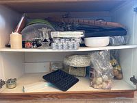 Wide view showing items stored on wooden shelf including candles, glass dish covers, a waffle maker plate, food storage containers and faux floral.