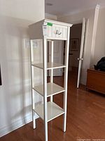 Front view of tall white shelving unit showing three open shelves and drawer with a metal hook attached.