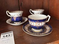 Three delicate teacups with matching saucers displayed on a wooden shelf, showcasing the gold and blue ornate pattern and elegant shapes.