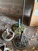 Three metal umbrella stands on concrete floor showing different decorative cast iron bases in green and black with visible rust and weathering.