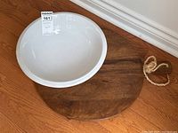 Photo showing white ironstone wash bowl sitting on round wooden cutting board with rope handle, placed on hardwood floor near white baseboard.