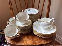 Photo showing stacked white porcelain Wedgwood tea cups with double handles, saucers, and small plates with gold trim stacked on wooden chair
