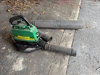 Full view of green and black Weed Eater blower/vac with two detachable black tubes on concrete and wood surface.
