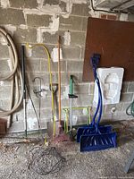 Yard tools including two blue plastic Yukon shovels, shepherd's hooks, metal shovel, rakes, and handheld cultivator on garage floor
