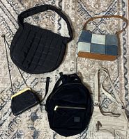 Top-down view of five bags on carpet: black quilted shoulder handbag, patchwork fabric shoulder handbag, black mesh small coin purse, black velvet small backpack, beige canvas shoulder bag.