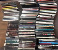 Multiple stacks of music CDs showing various artist names and album titles on spines, arranged horizontally and vertically in clear plastic jewel cases.