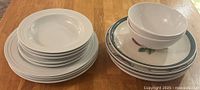 Assorted dinnerware including white plates and bowls stacked alongside decorative floral plates and bowl with green rim.