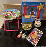 Photo showing all items in the lot positioned on a black surface, including the folding seat, coin bank, toothbrush holder, two keychains, and VHS tape.