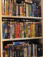 Top three shelves of a shelf filled with mostly VHS tapes and some DVDs of children's and family movies, with colorful covers.