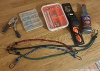Full lot overview showing bungee cords, power steering fluid, plastic organizers with nails and hardware, ratchet strap, and locking pliers on wood surface.