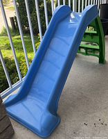 Side view of the blue plastic slide showing the entire sliding surface and green ladder support structure. Outdoor setting with shadows visible.