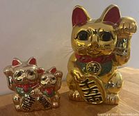 Three gold-colored Maneki Neko (beckoning cat) coin banks, one large and two smaller cats, displayed on a wooden surface showing front details.
