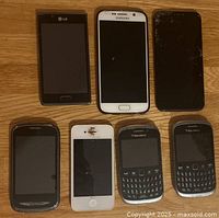 Seven mobile phones placed on a wooden surface showing various conditions of the front screens and keyboards. Phones include brands LG, Samsung, Apple, and BlackBerry.