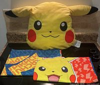 Yellow Pikachu-shaped pillow with two standard pillowcases with Pikachu-themed designs laid flat on countertop.