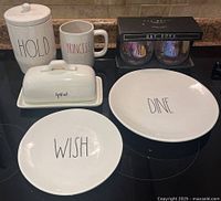Photo showing white ceramic jar with lid labeled 'HOLD', butter dish labeled 'spmad', mugs labeled 'PRINCESS' and 'TO THE MOON', plates labeled 'WISH' and 'DINE', and boxed glass cups.