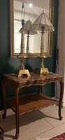 View showing wood side table with curved legs, scalloped lower shelf, two tall brass lamps with scalloped cream stained glass shades in front of a mirror.