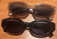 Two pairs of ladies sunglasses on a wooden surface. One pair features rectangular brown translucent frames and the other pair has black oval frames.