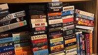 Photo shows various VHS tapes stacked on a shelf with visible titles including movies and TV shows.