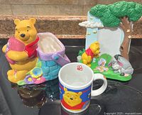 Photo showing entire lot of items including the Pooh planter, the photo frame with Pooh and Eeyore, and the Winnie the Pooh mug on a reflective black surface.