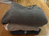 Three folded pieces of clothing stacked: grey knit hoodie with Michigan logo, beige top underneath, blue denim jeans at the bottom.