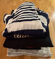 Stack of four folded women's clothing items including a navy and white striped top at the top, black top beneath, and grey striped fabric item at bottom.