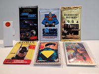 Photo of six sealed superhero collectible card packs arranged on a white surface with a small white stand showing '79'