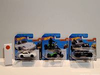 Three sealed Hot Wheels cars in original packaging: white Porsche, green off-road vehicle, and black pickup truck.