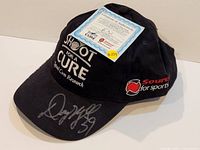 Black 'Shoot for a Cure' ball cap with silver signature on brim and Certificate of Authenticity on top of cap.