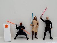 Three Star Wars figurines shown side by side against a plain white background. Characters from left to right: Darth Maul wielding double red lightsaber, Obi-Wan Kenobi with blue lightsaber, Count Dooku with red lightsaber raised above shoulder.