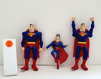Three Superman figurines in different poses, made of plastic, displayed against a white background with an orange #7 label to the left.
