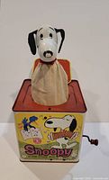 Full music box toy showing Snoopy head mounted on top with fabric skirt and red base tin box
