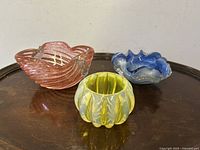 Three Italian mid-century modern glass bowls in yellow, pink, and blue, displayed together on a dark surface.