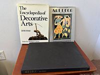 Three Art Deco themed books arranged standing and lying on wooden surface: The Encyclopedia of Decorative Arts 1890-1940, The All Color Book of Art Deco by Dan Klein, and Eva Weber's Art Deco hardcover without dust jacket.