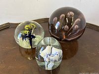 Three paperweights displayed on wooden surface. Large amethyst, medium yellow-black flower, small white flower with blue accents.