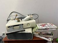 Two Xbox 360 consoles stacked, showing one with an open disc drive and power light illuminated. Two wired white controllers and a beige foam covered wired headset are on top. Stack of 7 Xbox 360 game cases including sports titles.