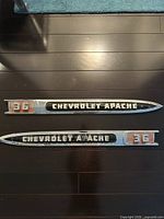 Pair of elongated chrome and painted 'CHEVROLET APACHE 36' emblems on dark surface, showing both emblems in good original condition with detailing.