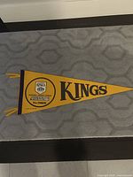 Full view of yellow and purple L.A. Kings hockey pennant laid flat on carpeted floor, showing large Kings text and logo with streamers.