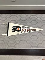 Full view of the white felt Philadelphia Flyers pennant laid flat showing entire design, including black and orange logo, word 'Flyers', black binding on wide end, and attached white streamers.
