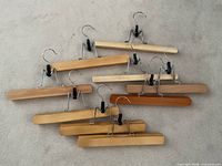 Photo showing nine wooden clamp hangers in various light to medium wood tones with metal hooks and black plastic clamp grips laid on a light surface.